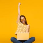 Young woman in yellow striped top with laptop raises arm in victory