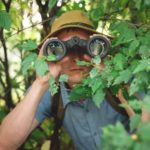 A man with binoculars crouched in the bush, indication a share price on watch