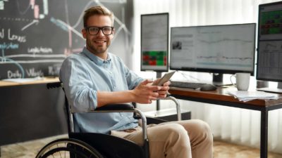 Smiling man with phone in wheelchair watching stocks and trends on computer