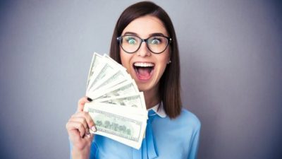 A happy woman wearing glasses and smiling broadly holds up a bunch of dollar notes