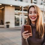 A happy woman stands outside a building looking at her phone and smiling widely