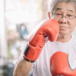 an elderly woman wearing boxing gloves raises one toward her face in a boxing pose while looking towards the camera with grey hair and spectacles on.