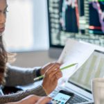 young woman reviewing financial reports at desk with multiple computer screens