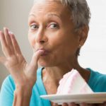 A mature woman holds a plate of cake and licks her thumb.