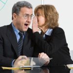 Businesswoman whispering in male colleague's ear as he looks surprised