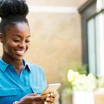 A woman standing in a blue shirt smiles as she uses her mobile phone to text message someone