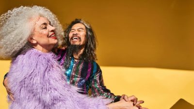 An older couple with wild hair in colourful clothes enjoy a moment dancing.