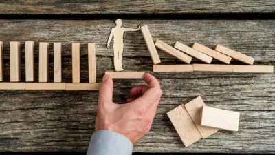 A hand holds a wooden figure up to a set of blocks to stop them falling, indicating life insurance policy