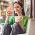 A smiling woman sits in a cafe reading a story on her phone about Rio Tinto and drinking a coffee with a laptop open in front of her.