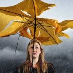 A woman with a sad face stands under a shredded umbrella in a grey thunderstorm