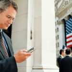 a business person checks his mobile phone outside a Wall Street office with an American flag and other business people in the background.