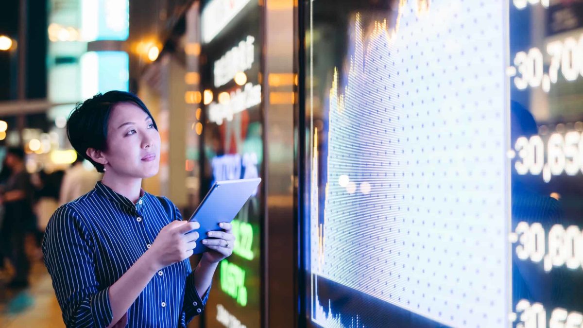 watch, woman with device standing next to large screen displaying rising share price information