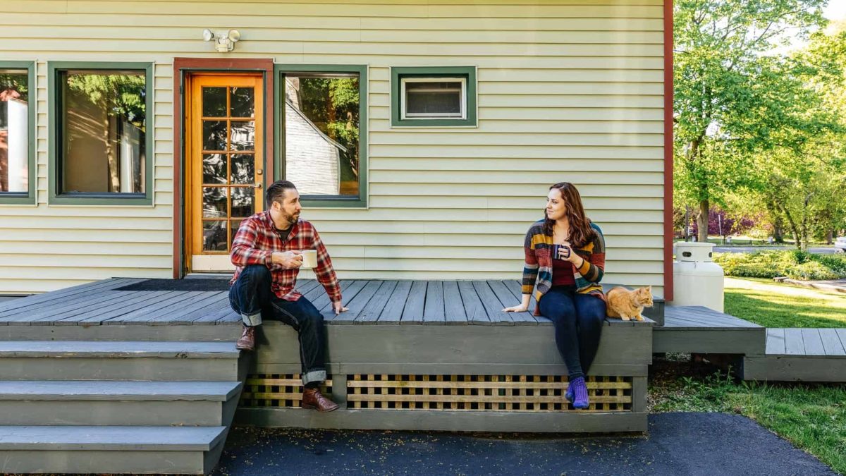 A man and a woman sit at either end of the front deck outside a house, looking at each other.