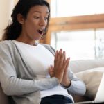 A young woman sits on her lounge looking pleasantly surprised at what she's seeing on her laptop screen as she reads about the South32 share price