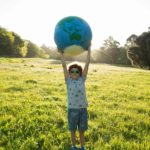 A young boy standing in a grassy field surrounded by trees holding a world globe over his head.