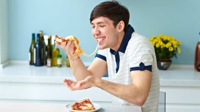 a happy man eats pizza in his kitchen with a long string of cheese between the pizza slice in his hand and in his mouth.