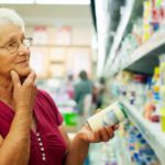 a woman ponders products on a supermarket shelf while holding a tin in one hand and holding her chin with the other.