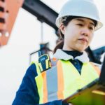 Female miner in hard hat and safety vest on laptop with mining drill in background.