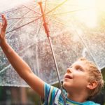 A young boy reaches up to touch the raindrops on his umbrella, as the sun comes out in the sky behind him.