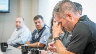 Man sits at table in office with head in hand as colleagues watch on