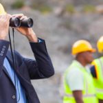 Pilbara Minerals engineer with hard hat looks through binoculars at work site or mine as two workers look on