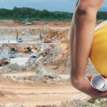 A miner holding a hard hat stands in the foreground of an open cut mine