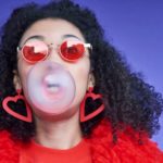 a woman wearing red blows a big bubble with bubblegum from her mouth.