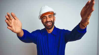 a man in a hard hat and overalls raises his arms and holds them out wide as he smiles widely in an optimistic and welcoming gesture.