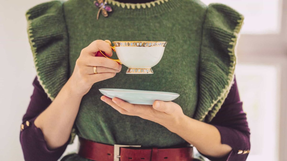 a woman holds a fine porcelain cup and saucer at chest level, raising the cup as though going to take a sip of tea.