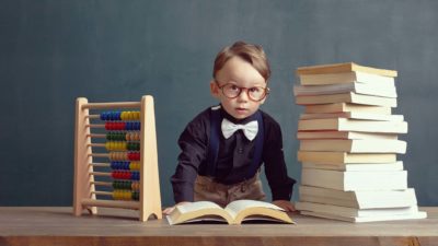 a small boy dressed in a bow tie and britches looks up from a pile of books with a book laid in front of him on a desk and an abacus on the other side, as though he is an accountant scouring books of figures.