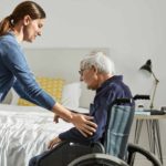 A young female nurse assists a male elderly resident at an aged care home get out of his wheelchair