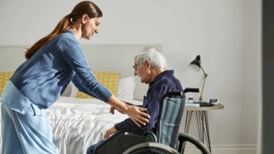 A young female nurse assists a male elderly resident at an aged care home get out of his wheelchair