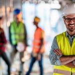 Male building supervisor wearing high vis vest and hard hat stands and smiles with his arms crossed at a building site