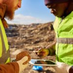 Two miners dressed in hard hats and high vis gear standing at an outdoor mining site discussing a mineral find with one holding a rock and the other looking at a tablet.