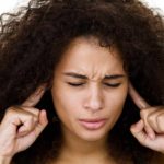 a woman puts her fingers in her ears with a pained expression on her face with her eyes closed as though trying to block hearing bad news or an unpleasant loud noise.