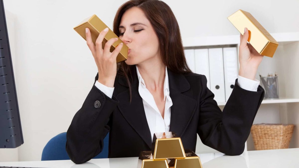 A woman in a business suit sits at her desk with gold bars in each hand while she kisses one bar with her eyes closed. Her desk has another three gold bars stacked in front of her. symbolising the rising Northern Star share price