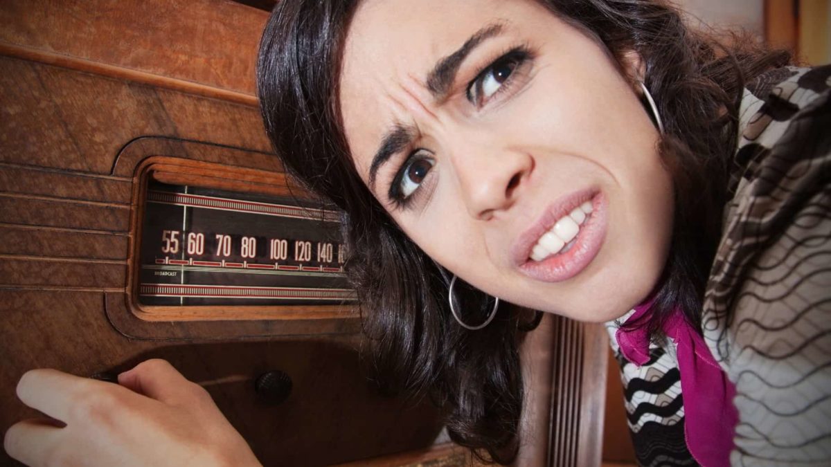 a woman with a pained and confused look on her face twiddles the dial on an old fashioned radio receiver while leaning in to listen to it.