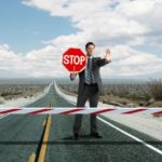 a man in a suit holds up a hand and a stop sign at a roadblock positioned over a bitumen road .