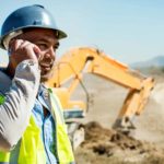 A man in a hard hat and high visibility vest speaks on his mobile phone in front of a digging machine with a heavy dump truck vehicle also visible in the background.