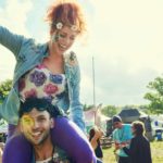 a woman wearing a flower garland sits atop the shoulders of a man celebrating a happy time in the outdoors with people talking in groups in the background, perhaps at an outdoor markets or music festival, in an image portraying young people enjoying freedom.