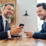 Two laughing male executives wearing dark suits chat across a timber lunch room table while one of them holds up his phone to show information.