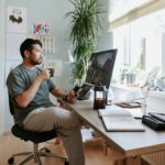 A young man working from home sits at his home office desk holding a cup of tea and looking out the window