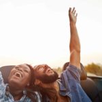 A young couple in the back of a convertible car each raise a single arm in the air whilst enjoying a drive along the road.