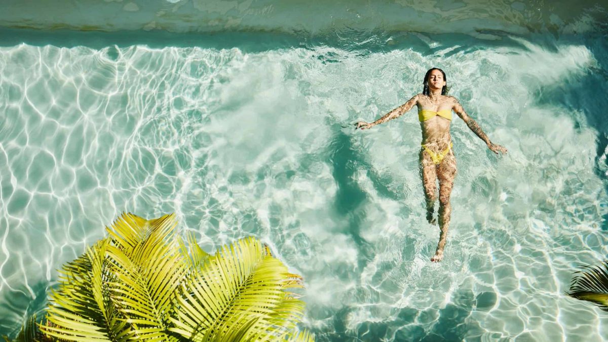 An overhead aerial photo of a young woman swimming in a hotel pool wearing a yellow bikini on holiday