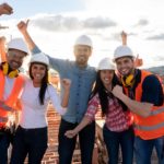 a group of five engineers wearing hard hats and some in high visibility vests raise their arms in happy celebration atop a building site with construction and equipment in the background.