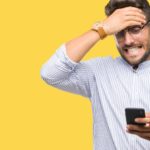 a man clasps his hand to his forehead as he looks down at his phone and grimaces with a pained expression on his face as he watches the Pilbara Minerals share price continue to fall