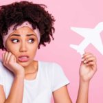 A woman looks nervous and uncertain holding a hand to her chin while looking at a paper cut out of a plane that she's holding in her other hand. representing the falling Air New Zealand share price today