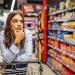 A female Woolworths customer leans on her shopping trolley as she rests her chin in her hand thinking about what to buy for dinner while also wondering why the Woolworths share price isn't doing as well as Coles recently