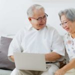 an older couple look happy as they sit at a laptop computer in their home.