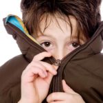 a boy with sad eyes pulls the zip over his mouth and nose while doing up a large jacket where the collar stands up at head height.